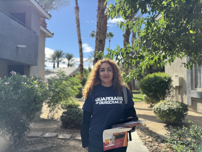 woman smiling, with dark long curly hair wearing a guardians of democracy shirt, holding a stack of papers