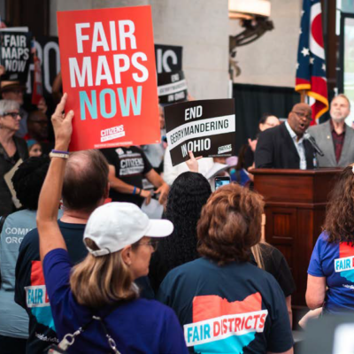 A man with dark skin speaking to a large group of people, some are holding signs. One sign says Fair maps now.