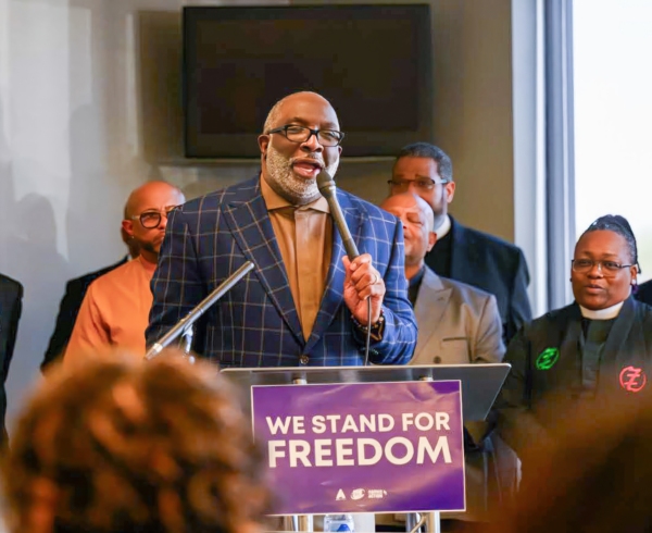 A man with dark skin and a beard standing at a lectern, speaking into a microphone. A sign that says We Stand for Freedom is hanging on the lectern. Several people stand behind him.