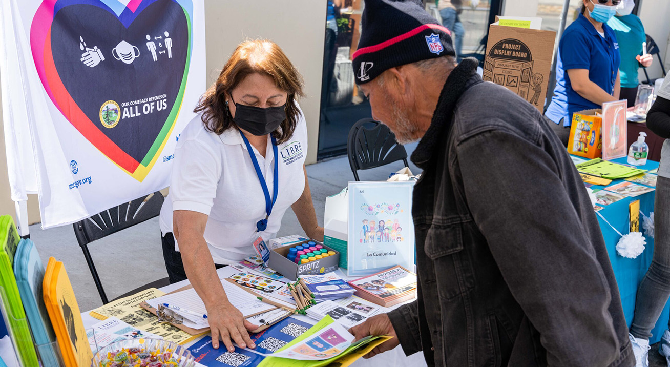 A woman at a table at an outdoor event shows a man some brochures.